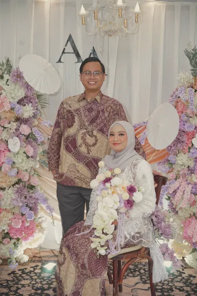 Andrika berdiri di belakang Aliva yang duduk memegang buket, di depan latar bunga.