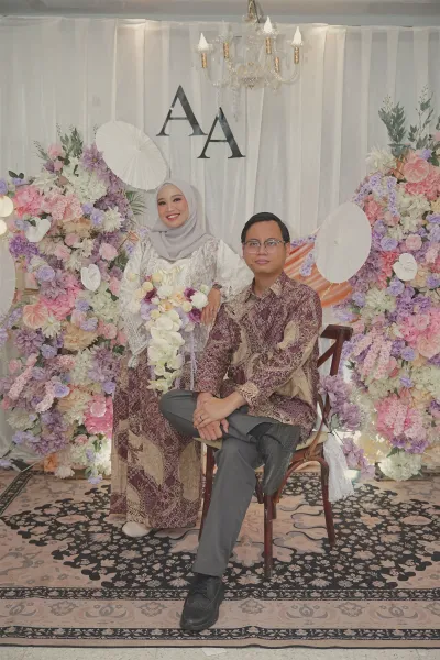 Aliva berdiri di belakang Andrika yang duduk di kursi kayu, di depan latar bunga.