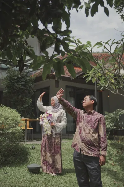 Andrika mengangkat tangan sambil tertawa bersama Aliva di taman.
